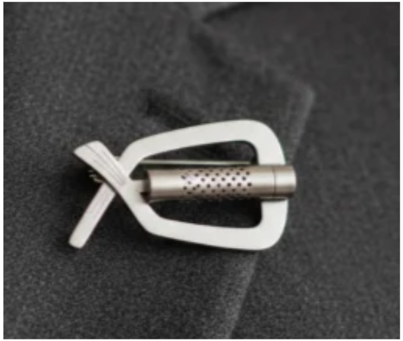 Broche parfumable Osmélya en acier inoxydable argenté, fixée sur un vêtement en tissu gris, avec capsule textile rechargeable intégrée